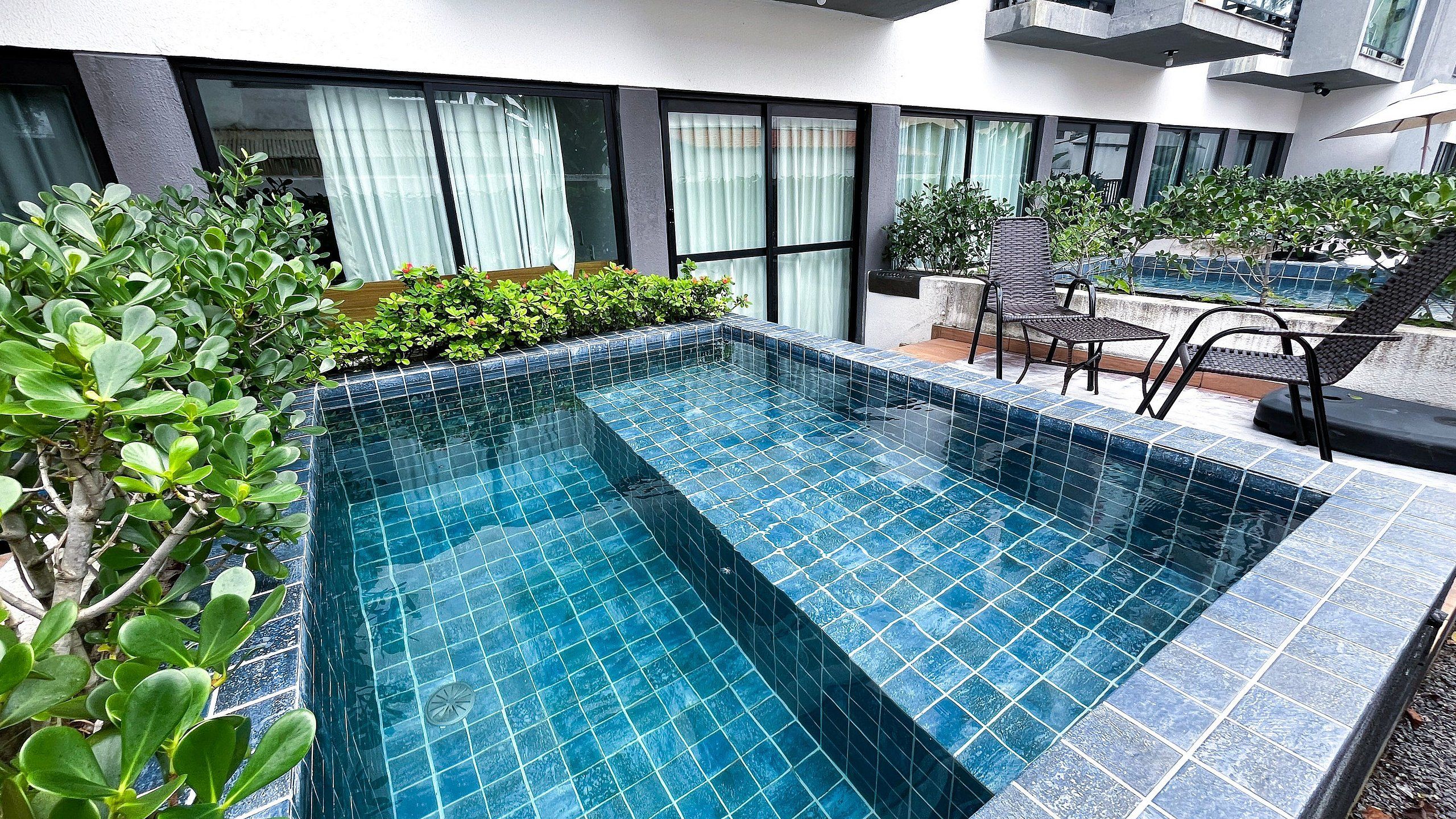 A rectangular outdoor swimming pool with blue tiles, surrounded by greenery and modern outdoor furniture.