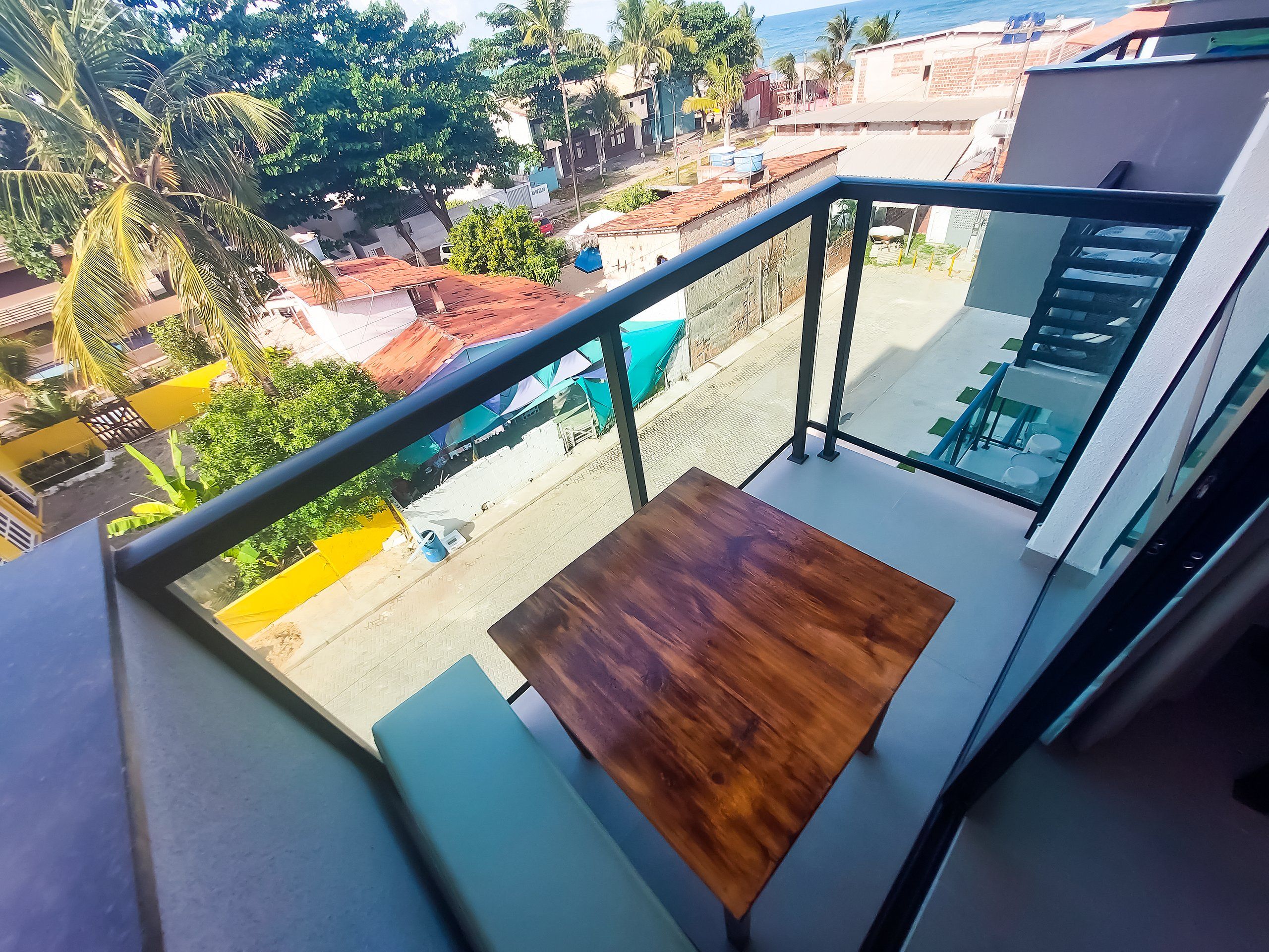 Ein Balkon mit Holztisch, Metallgeländer und Blick auf eine Straße mit Gebäuden und Palmen.
