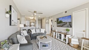 A modern living room with a grey sofa, a coffee table, and a TV. There is a kitchen in the background with white cabinets and a dining area with a wooden table. - Ocean Links - Stunning 3rd Floor Ocean View! (Hilton Head)