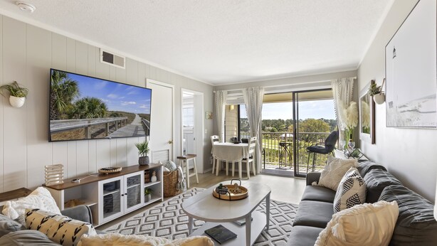 A spacious living room with a large flat-screen TV, a sofa, a coffee table, and a balcony with a view of greenery. - Ocean Links - Stunning 3rd Floor Ocean View! (Hilton Head)
