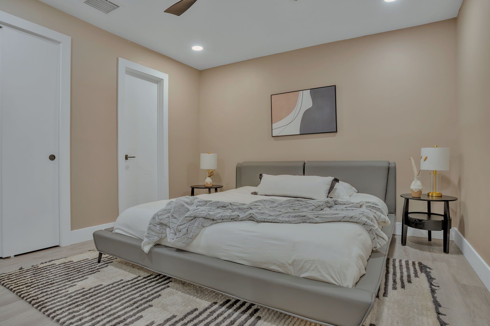 A bedroom with a bed, two bedside tables with lamps, a door, and a wall-mounted artwork.