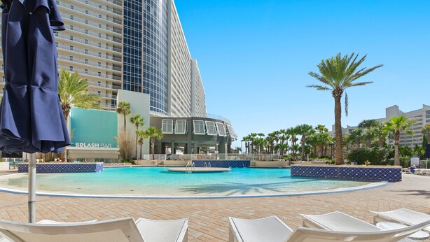 A pool area with lounge chairs, a palm tree, and a building with a sign reading "SPLASH BAR". - Gulf views! Spacious with 2 King beds, Bunk Room | 5 Pools (Panama City Beach)