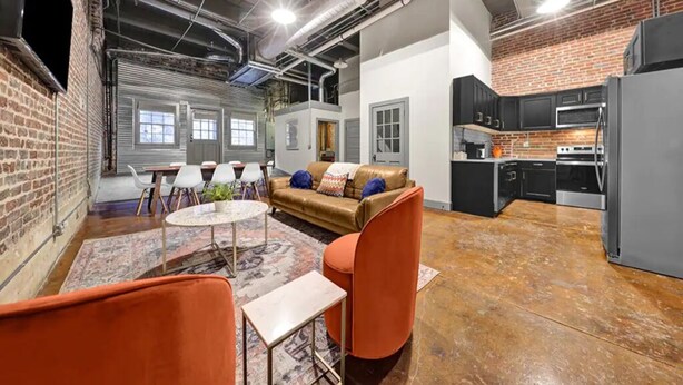 A spacious loft with a high ceiling, exposed ductwork, a brick wall, a cozy seating area with a sofa and chairs, and a kitchenette with modern appliances. - Penthouse Loft Downtown Huge Unit (Birmingham)