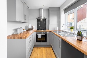 A modern kitchen with grey cabinets, a wooden countertop, a built-in oven, a stainless steel range hood, and a window with a view of the street.