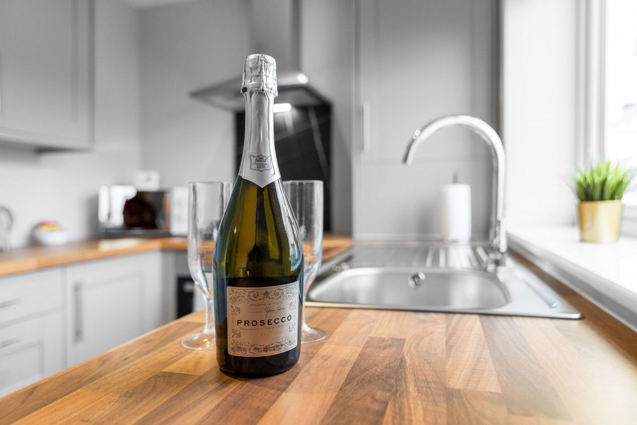 A bottle of Prosecco wine on a wooden countertop in a modern kitchen with a stainless steel sink and a potted plant.