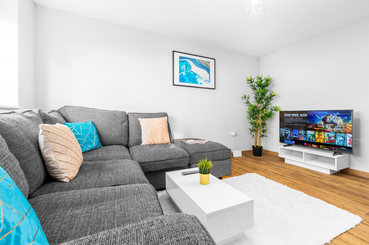Un salon moderne avec un canapé d’angle gris, une table basse blanche, un téléviseur à écran plat et une plante en pot.