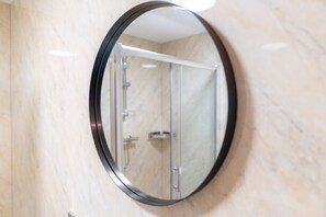 A round mirror with a black frame reflecting a bathroom with a glass shower enclosure and a marble wall.