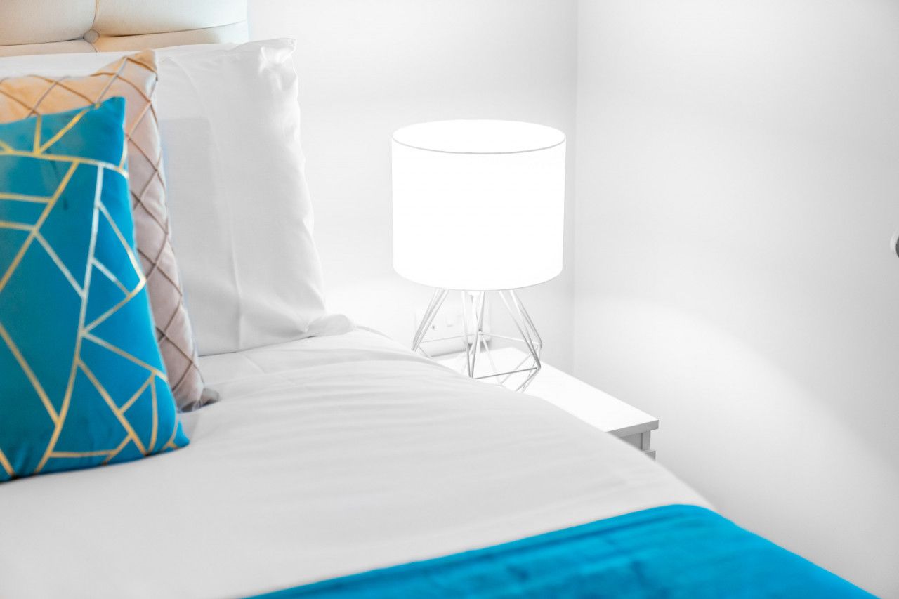 Una cama bien hecha con ropa blanca y una manta de color azul, una almohada estampada con motivos geométricos y una lámpara de mesa moderna sobre una mesita de noche.