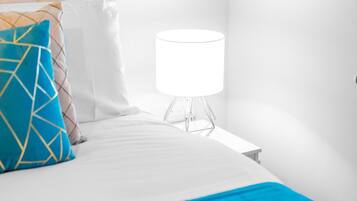 Una cama bien hecha con ropa blanca y una manta de color azul, una almohada estampada con motivos geométricos y una lámpara de mesa moderna sobre una mesita de noche.