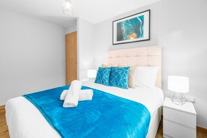 Moderni hotellihuone, jossa on suuri sänky, valkoinen yöpöytä, seinällä oleva taideteos kehyksissä ja puinen ovi.