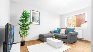 En moderne stue med en grå sofa, et hvidt sofabord, et stort fladskærms-TV, en potteplante og et vindue med udsigt til en murstensbygning.