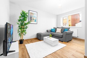 A modern living room with a grey sofa, a white coffee table, a large potted plant, and a flat-screen TV. - Stylish Two Bed - Two Parking Spaces - Stourbridge (West Midlands)