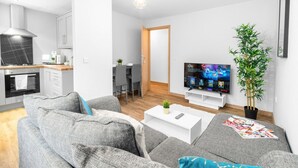 En moderne stue med en grå sofa, et sofabord og et fjernsyn, der viser et quizprogram. I baggrunden ses et spiseområde med et træbord og stole.