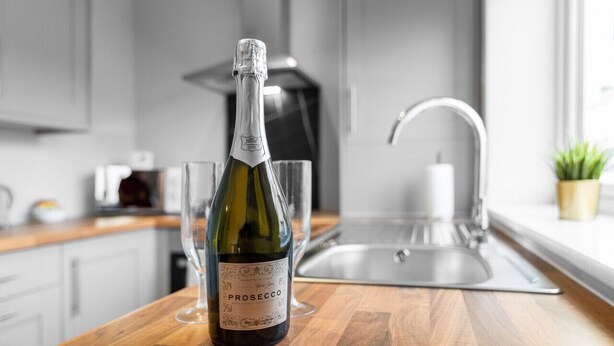 A bottle of Prosecco wine on a wooden countertop in a modern kitchen with a stainless steel sink and a potted plant. - Brand New Apartment - Parking - Stourbridge (Stourbridge)