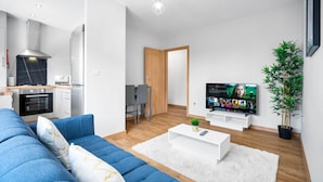 Una sala de estar moderna con un sofá azul, una mesa de centro blanca y un televisor que muestra una película. Se ve una cocina con estufa y armarios al fondo.