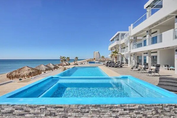 A swimming pool with lounge chairs and a beach view, surrounded by a building with balconies.