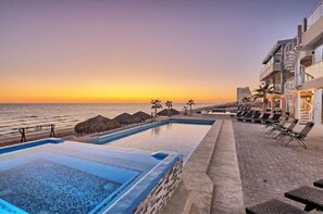 Un espace piscine avec vue sur l’océan au coucher du soleil, des chaises longues et un bâtiment doté de balcons.