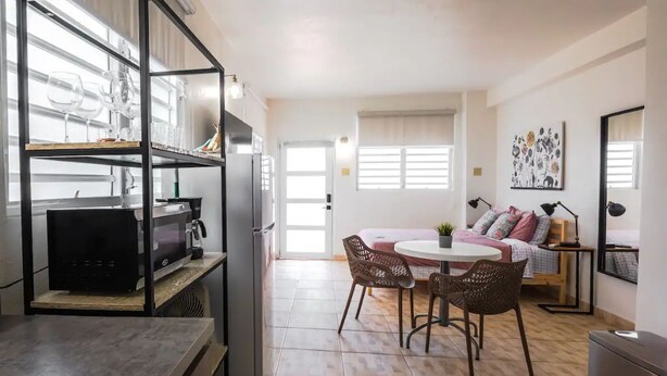 A compact living space with a kitchenette, a dining area with wicker chairs, a bed with a floral bedspread, and a window with a view. - City Park View - Cozy Studio (Guaynabo)