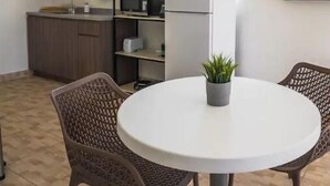 A compact kitchen with a refrigerator, microwave, and shelves. There is a round table with chairs and a potted plant. - City Park View - Cozy Studio (Guaynabo)