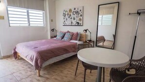 A bedroom with a bed, a round table, two chairs, a nightstand, a lamp, a mirror, and a floral artwork on the wall. - City Park View - Cozy Studio (Guaynabo)