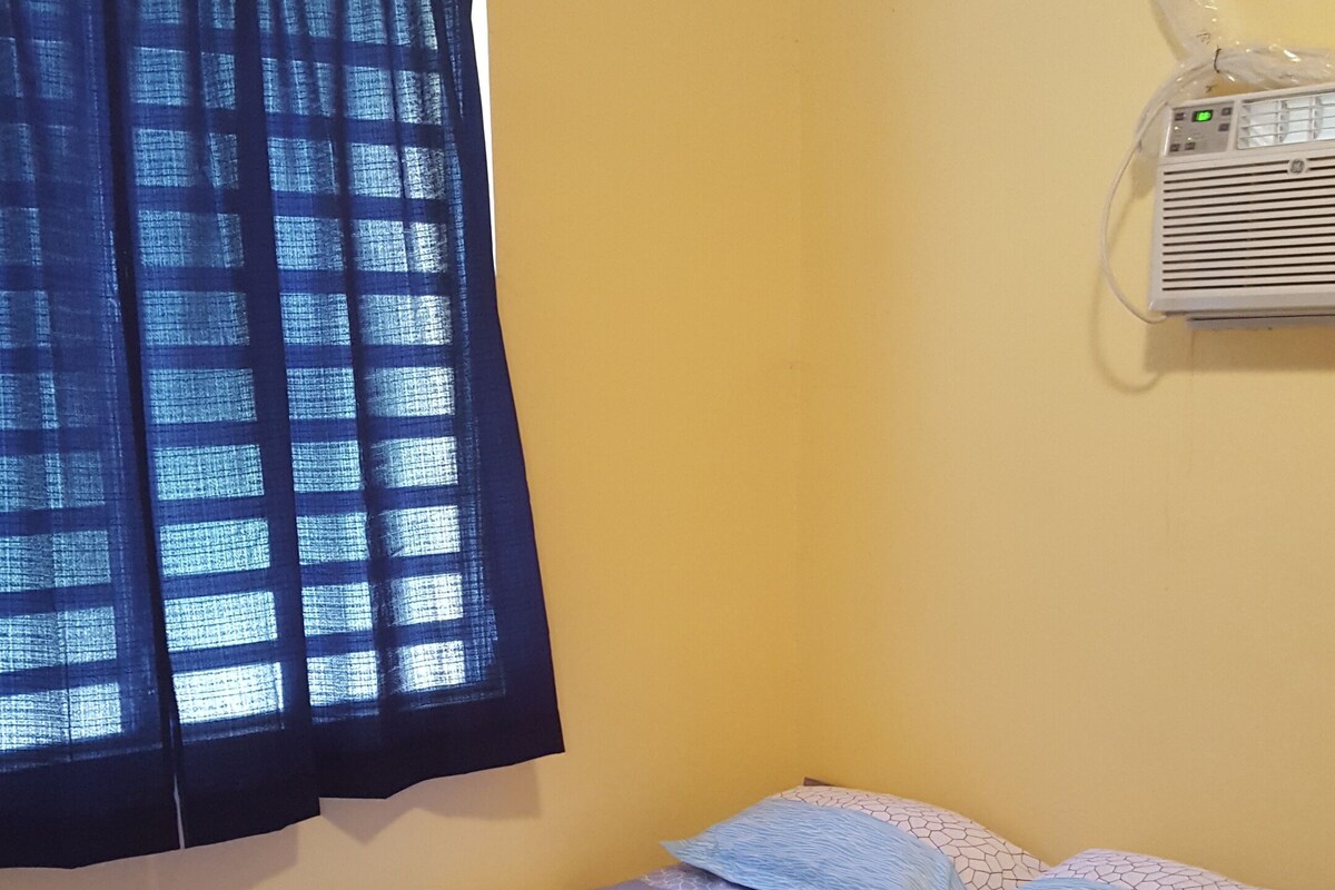 A small, single-bed room with a plain yellow wall, a window with blue curtains, and a wall-mounted air conditioner.