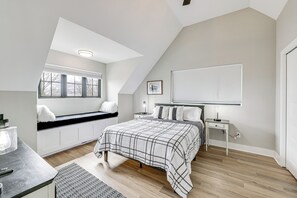 Un dormitorio con techo inclinado, una cama con colcha escocesa, un rincón con asiento junto a la ventana, una mesita de noche con lámpara y otra mesita auxiliar.