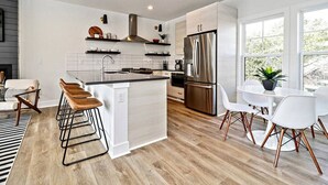 A modern kitchen with a central island, stainless steel appliances, and a dining area with white chairs. - Rooftop Deck and Ocean Views with VB Local Life! (Virginia Beach)