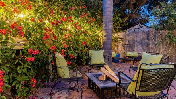 Un patio exterior con un brasero y sillas, rodeado de vegetación exuberante y plantas con flores.