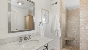 Un baño con lavamanos de mármol, un espejo grande, una impresión botánica enmarco y una zona de ducha con azulejos color beige.