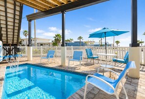 A swimming pool with blue chairs and umbrellas, surrounded by a white fence and palm trees.