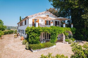 Et tosidet hus med en overdækket terrasse, en stenindhegnet gårdsplads og frodig vegetation.