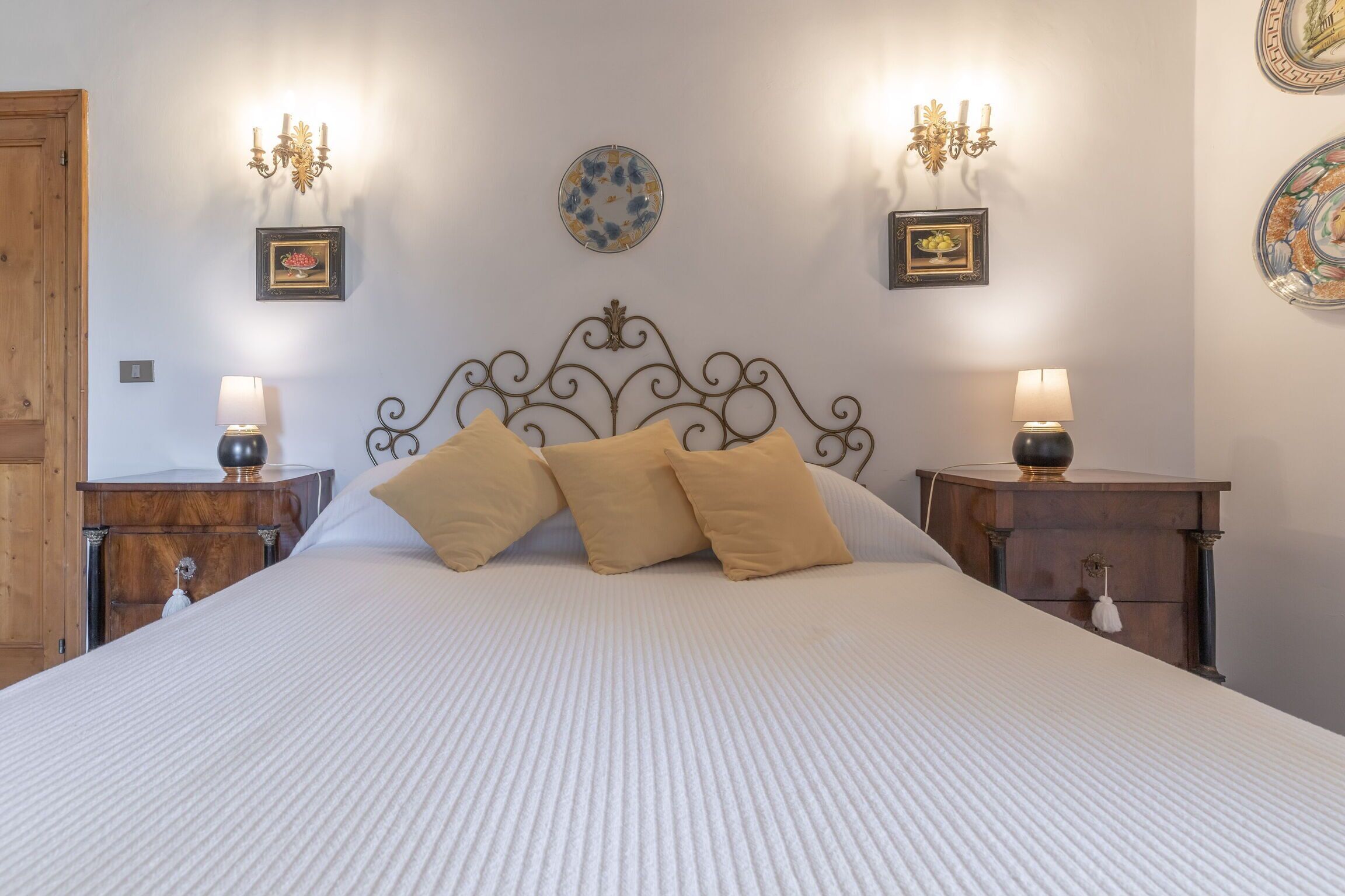 A neatly made bed with a white bedspread and three beige pillows, flanked by two bedside tables with lamps, a wall-mounted decorative plate, and a wooden door.