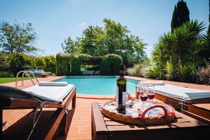 Et poolområde med liggestole, et bord med en flaske og glas, samt udsigt til poolen og den omkringliggende vegetation.