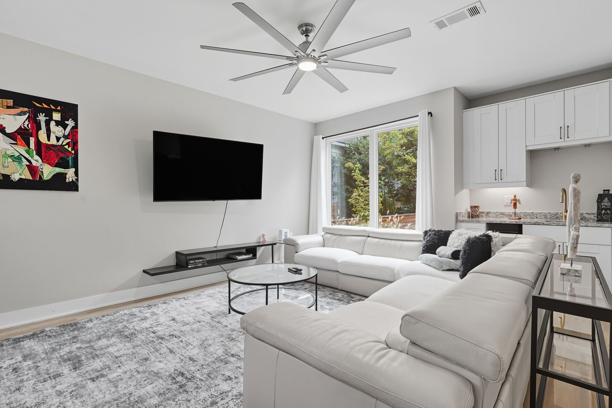 Un salon moderne doté d’un grand canapé d’angle, d’un téléviseur à écran plat fixé au mur, d’un ventilateur de plafond et d’une grande fenêtre donnant sur un paysage verdoyant.