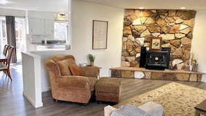 Una sala de estar con chimenea de piedra, un sofá, un tapete estampado y un comedor con una cocina al fondo.