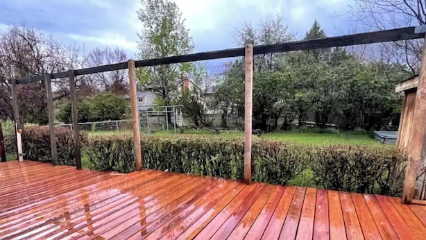 Una terraza de madera con barandilla de metal, rodeada de setos y árboles.