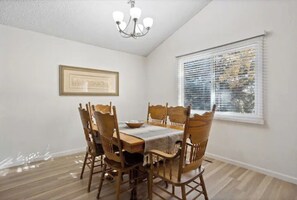 Une salle à manger avec une table et des chaises en bois, un lustre, un tableau encadré au mur et une fenêtre avec des stores.