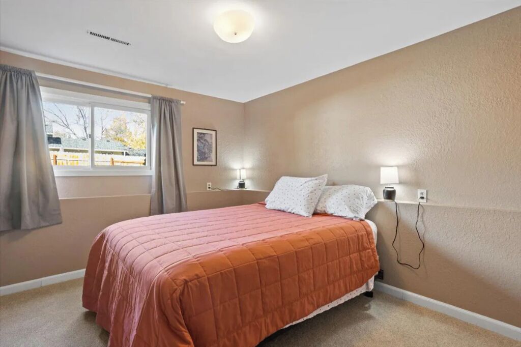 Une chambre à coucher avec un lit, deux lampes de chevet, une fenêtre avec des rideaux et une prise électrique fixée au mur.
