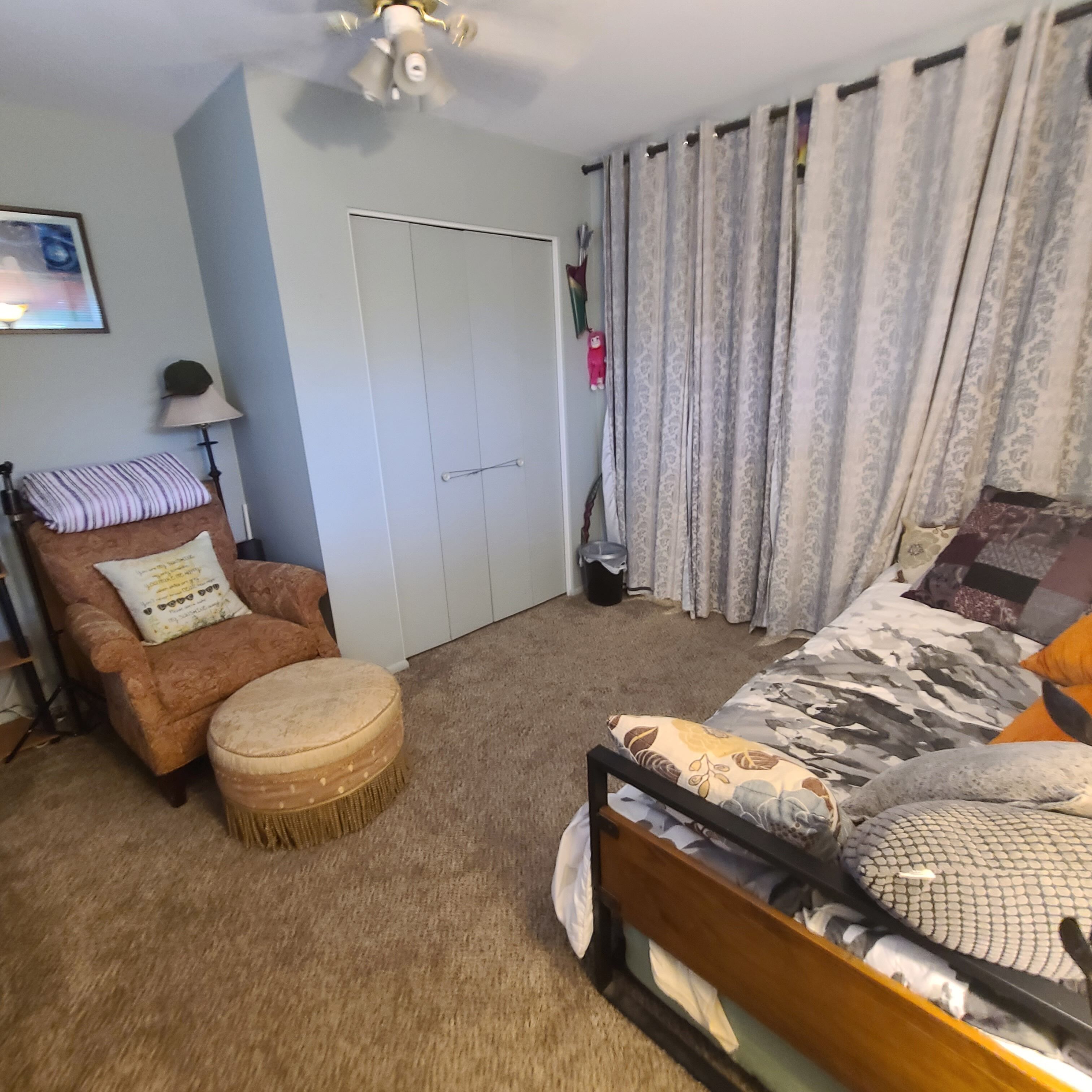 A bedroom with a bed, a chair, a small ottoman, a wardrobe, and a ceiling fan.