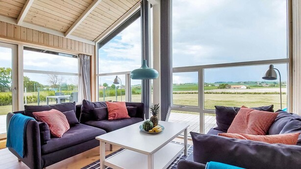 Un vaste espace de vie au design moderne, comprenant un ensemble de canapés aux couleurs sombres, une table basse blanche et de grandes fenêtres offrant une vue sur la campagne.