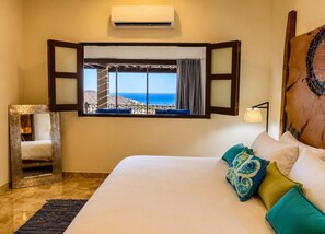 A hotel room with a large bed, a view of the sea, a window with curtains, and an air conditioning unit.