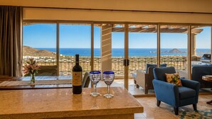 Una habitación con un ventanal grande, una mesa con una botella y vasos, una silla y vista al mar.