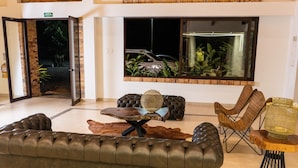 Un hall d’entrée avec un canapé en cuir, une table basse en bois et une grande fenêtre donnant sur une voiture et des plantes à l’extérieur.