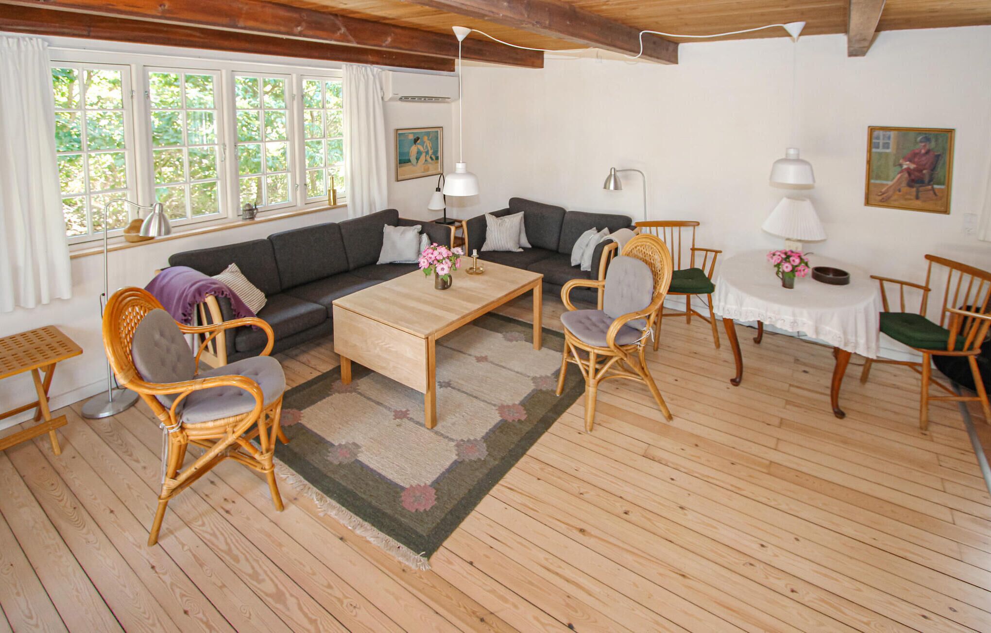 Una sala acogedora con un sofá, dos sillones, una mesita de centro y una mesa de comedor con sillas. Hay ventanas amplias con cortinas, un cuadro en la pared y una lámpara en la mesa.
