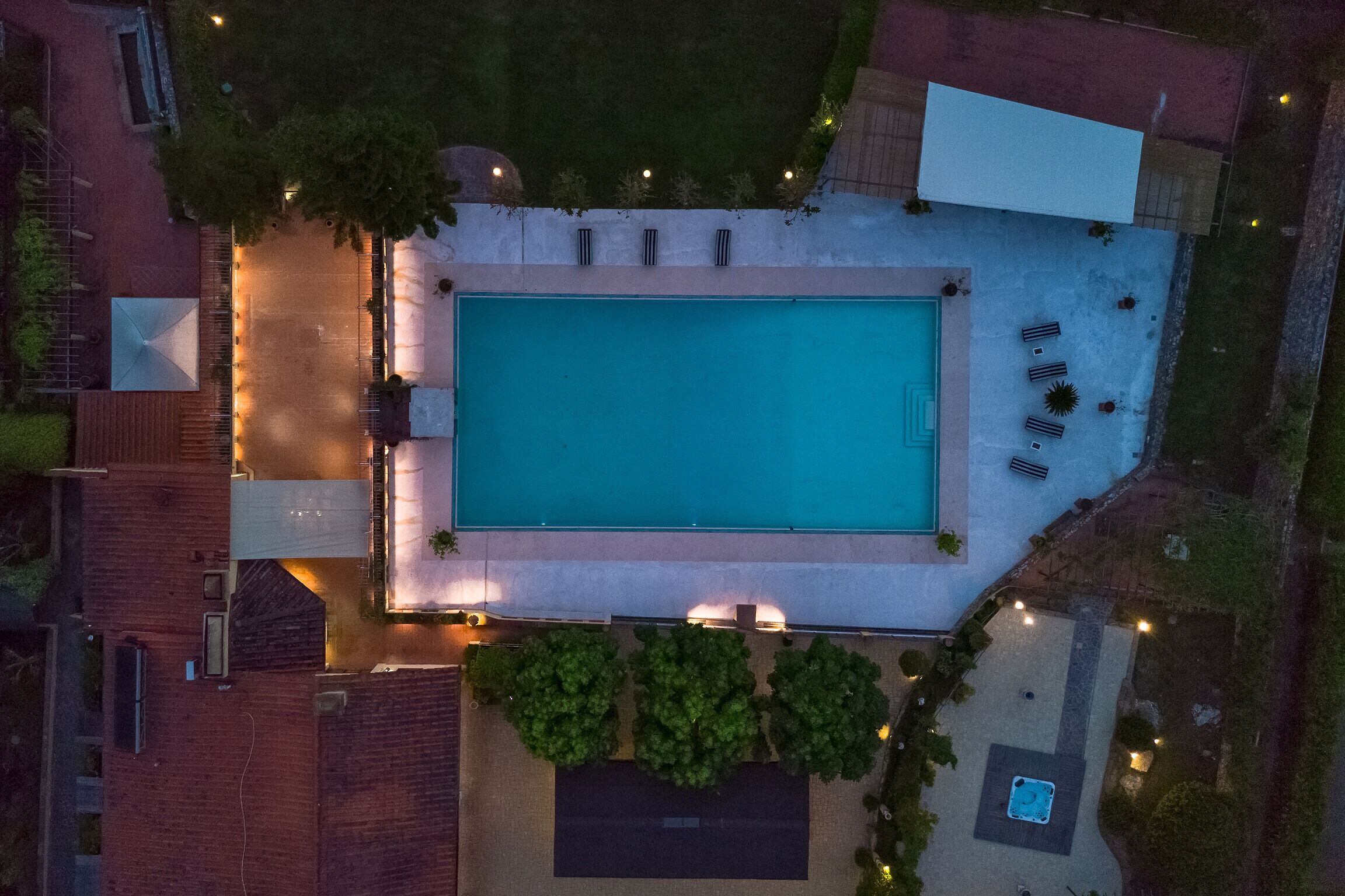Vue aérienne d’une piscine rectangulaire entourée d’une terrasse, d’un espace de détente couvert et d’un petit jardin agrémenté de plantes en pot.