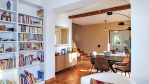 Een ruime kamer met een grote boekenkast vol boeken, een eethoek met een tafel en stoelen, en een trap naar een ander niveau.