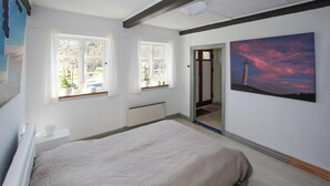 Un dormitorio con una cama, dos ventanas, un radiador, una puerta que da a otra habitación y un cuadro de un faro.