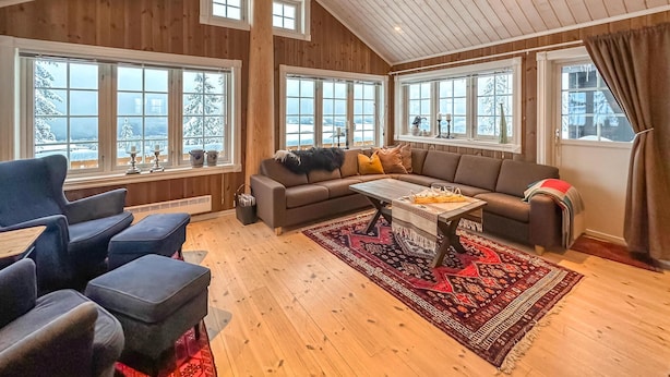 Een gezellige woonkamer met een ruime hoekbank, een salontafel en een rood tapijt met een patroon. Er zijn meerdere ramen met uitzicht op met sneeuw bedekte bomen.