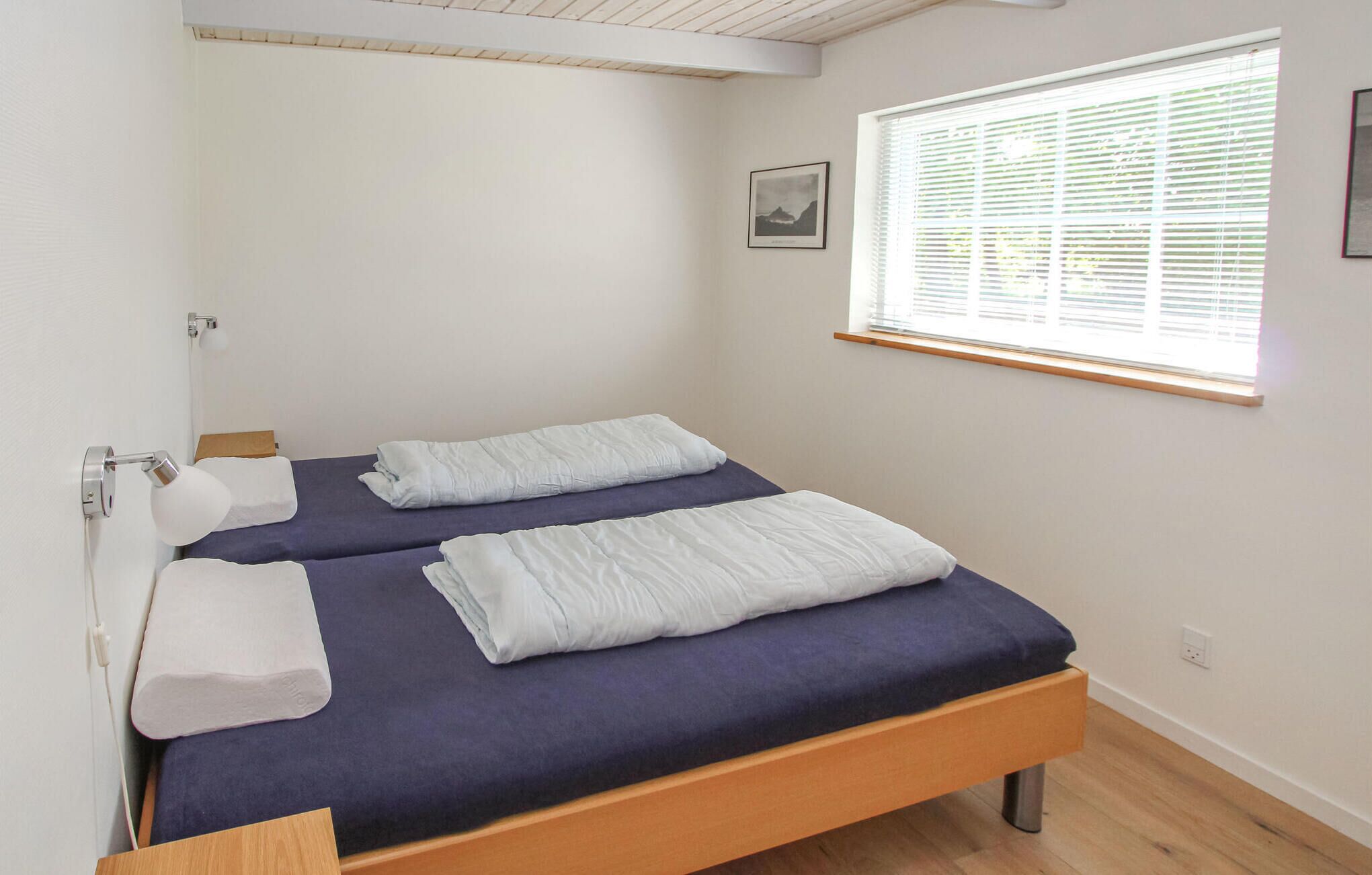 A small, neatly made bed with a blue mattress and white pillows, a wooden headboard, and a window with blinds.