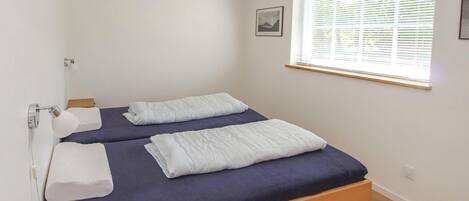 A small, neatly made bed with a blue mattress and white pillows, a wooden headboard, and a window with blinds.